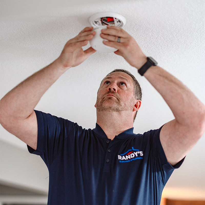A man in a blue uniform installs a smoke detector on a ceiling.