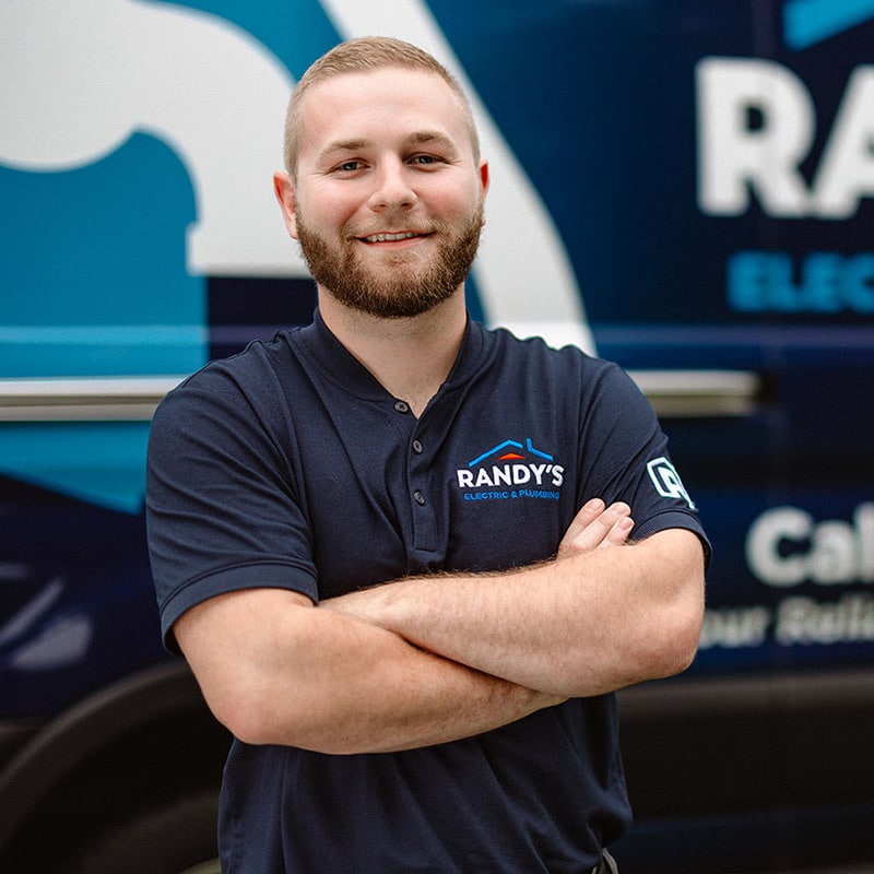 A man in a Randy's Electric & Plumbing uniform smiling with arms crossed, standing in front of a branded vehicle.